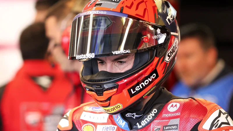 Marc Marquez mengenakan helm Shoei merah dan baju balap tim Ducati Lenovo, tampak menunduk fokus di area paddock MotoGP.