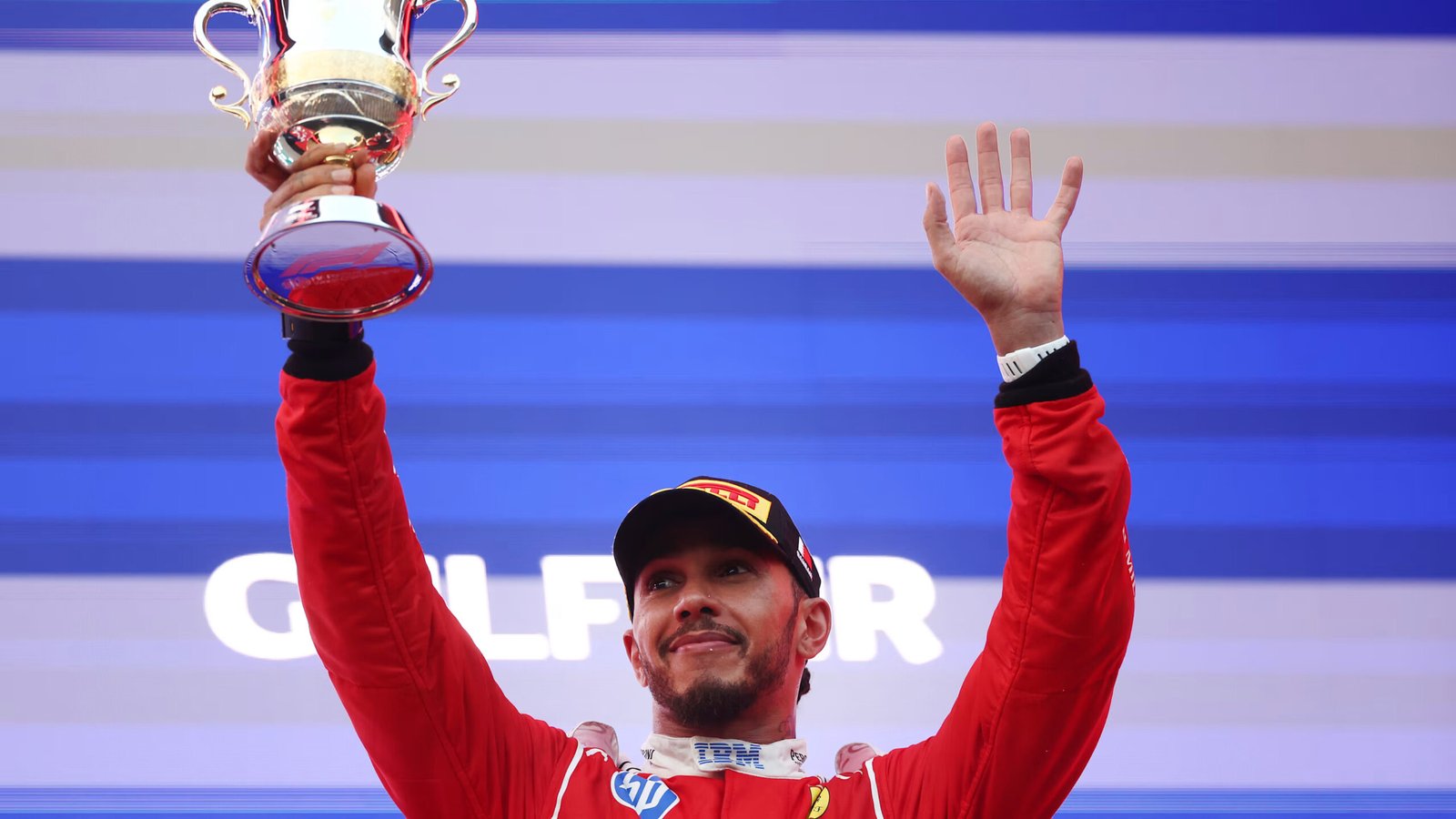Lewis Hamilton mengenakan seragam Ferrari, mengangkat trofi dan melambaikan tangan di podium Sepang.