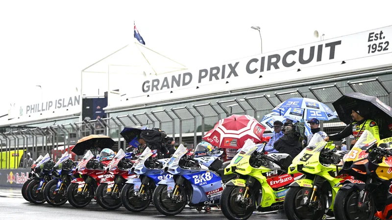 Deretan motor MotoGP dan kru dengan payung di lintasan basah Phillip Island.