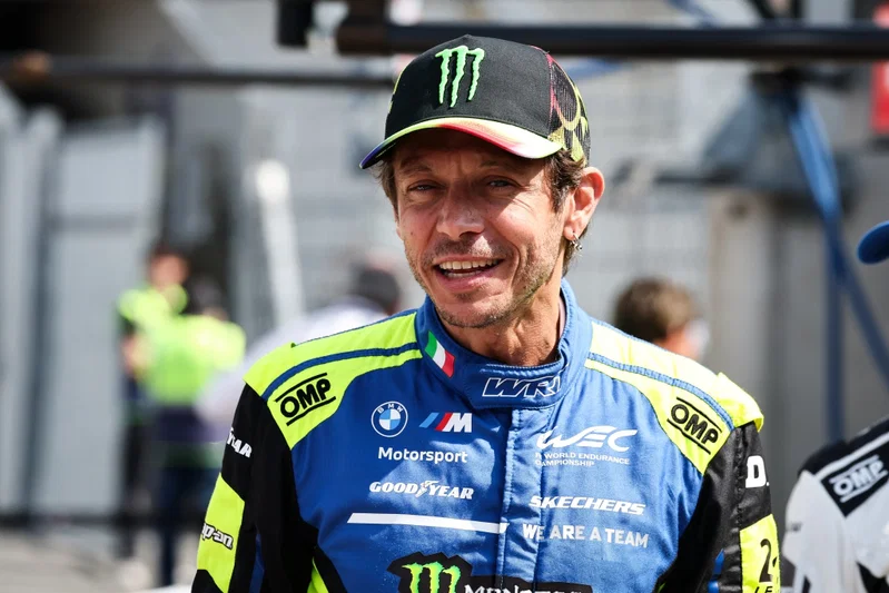 Valentino Rossi tersenyum mengenakan topi hitam dan baju balap biru Team WRT dengan detail kuning.