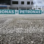 Detail permukaan aspal Sirkuit Sepang dengan tekstur kasar dan papan Petronas di latar belakang.