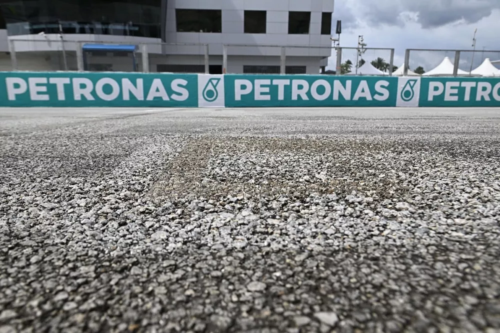 Detail permukaan aspal Sirkuit Sepang dengan tekstur kasar dan papan Petronas di latar belakang.